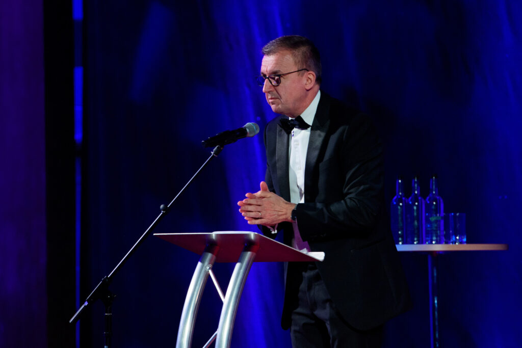 A person in a suit and bow tie speaks into a microphone on a stand, standing at a podium. The backdrop of the Lincolnshire Business Awards 2025 is dark blue, with a small table holding several bottles visible to the side.