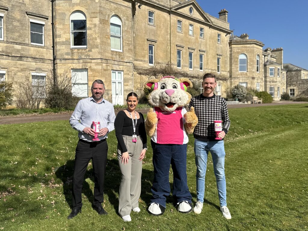 Four people, including a mascot in a pink outfit, stand on grass in front of a historic stone building. Two individuals hold Cash for Kids Charity donation buckets. The Waldeck Wellingore Team enjoys the clear and sunny sky as they prepare to support the Lincoln 10K event.