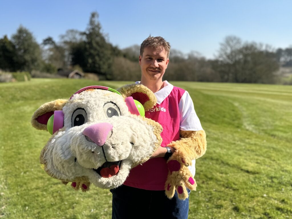 A person in a pink shirt stands on a grassy field, holding a large, smiling costume head of an animal with headphones, promoting Cash for Kids. Trees and the clear sky provide the perfect backdrop for this fun moment with the Waldeck Wellingore Team.